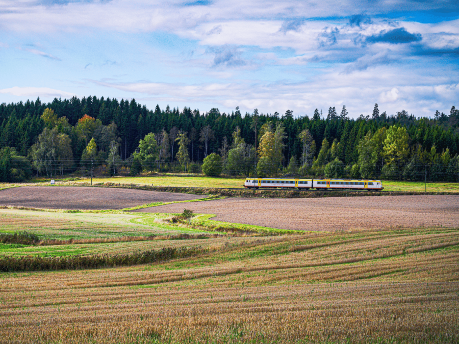 A regional passenger train travelling through open farmland with forest in the background under a partly cloudy sky.