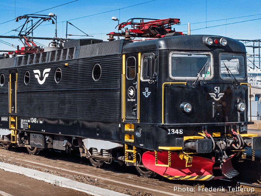 Schwarze SJ Rc6-Elektrolokomotive an einem schwedischen Bahnhof, mit Stromabnehmer und Hochspannungsausrüstung auf dem Dach.